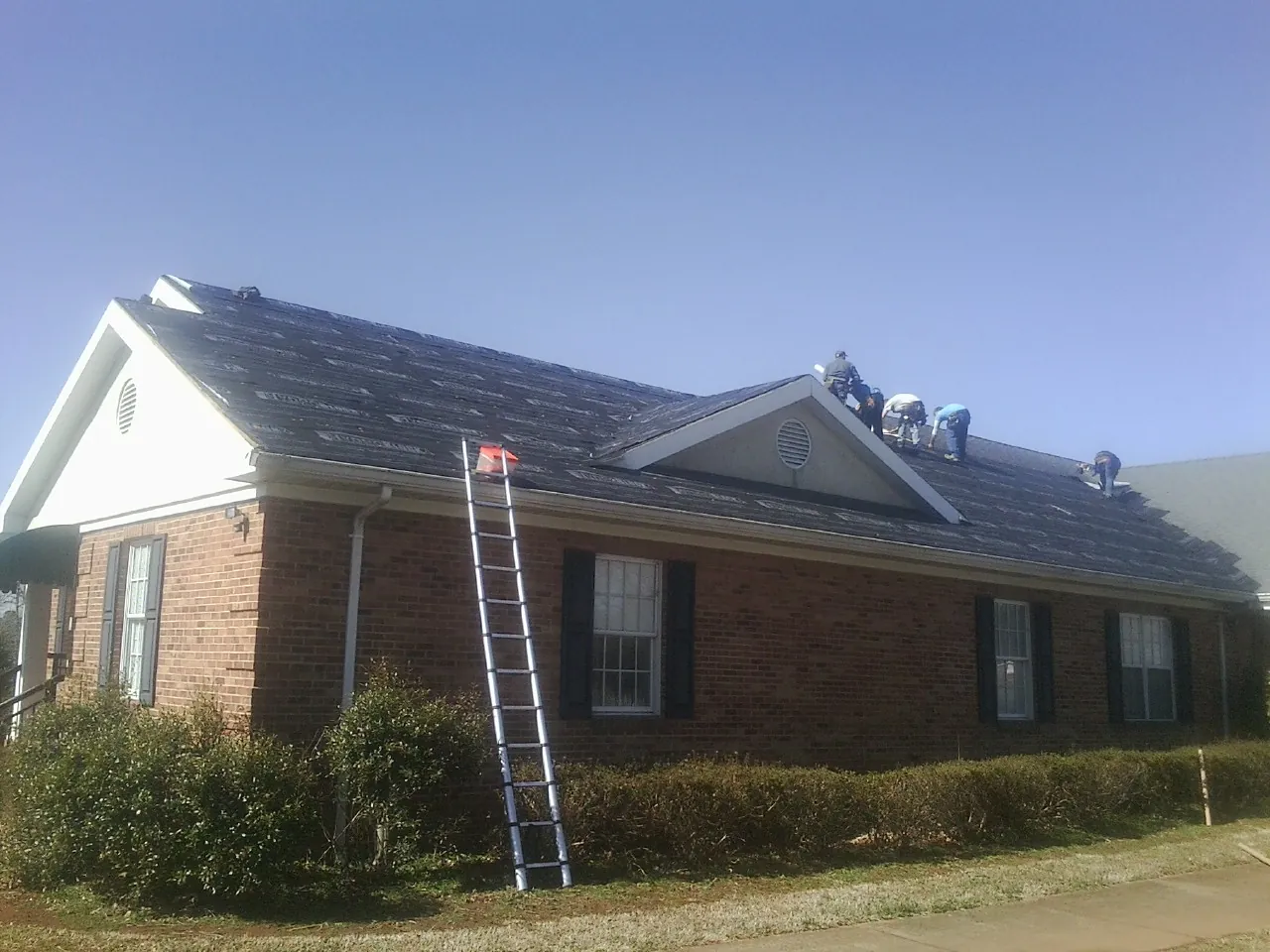 Crew tearing shingles off a brick ranch roof with a ladder set — Emergency Roof Repair work by our crew in West Warwick