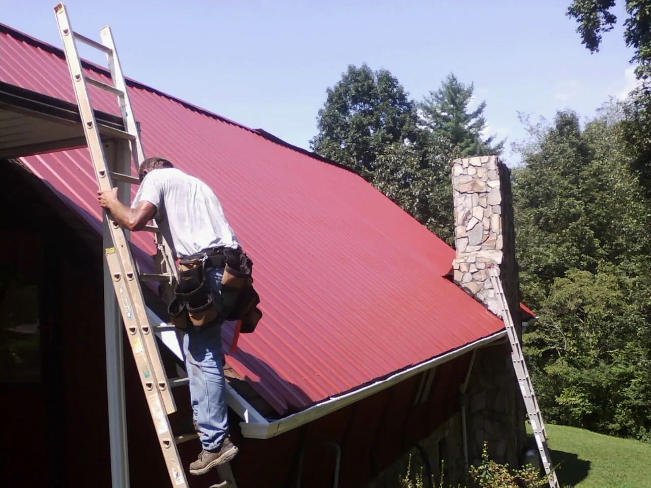 Roofer on a ladder beside a red metal roof and stone chimney — Soffit & Fascia Repair by Roofing Brothers in West Warwick